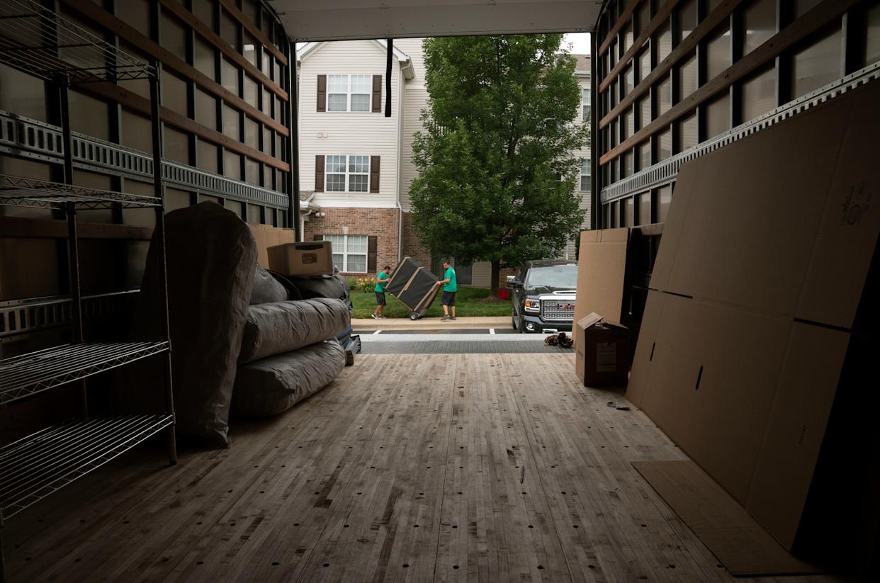 The alt text for the image is: Two men in green shirts moving furniture in a parking lot with a large truck in the backgro...